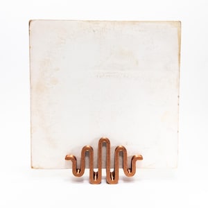 Puede incluir: Un tarjetero de metal color cobre con un diseño ondulado frente a un fondo rectangular blanco ligeramente envejecido. El tarjetero tiene una serie de bucles y curvas verticales.