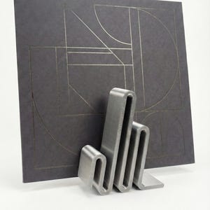May include: A dark gray album cover with a geometric design in silver is propped up by a silver metal stand. The stand has a unique, wavy design. The album cover features lines and shapes.