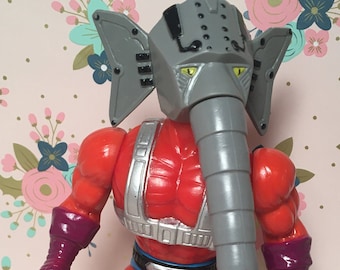Puede incluir: Una figura de acción roja y gris con cabeza de elefante y una larga trompa gris. La figura lleva una pieza de pecho plateada y un taparrabos azul.