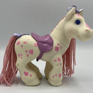 May include: A white toy horse with pink spots and a purple saddle. The horse has a pink yarn mane and tail.