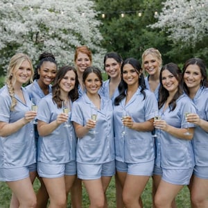 May include: A group of people wearing matching light blue pajama sets, holding champagne flutes. The pajama sets include short-sleeved button-down shirts and shorts. The background features a lush green lawn and white flowering trees.