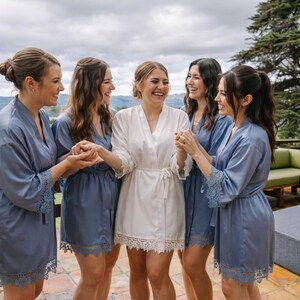 May include: A group of five individuals wearing matching robes. One individual in the center wears a white satin robe with lace trim, while the four surrounding individuals wear dusty blue satin robes with matching lace trim on the sleeves and hem.