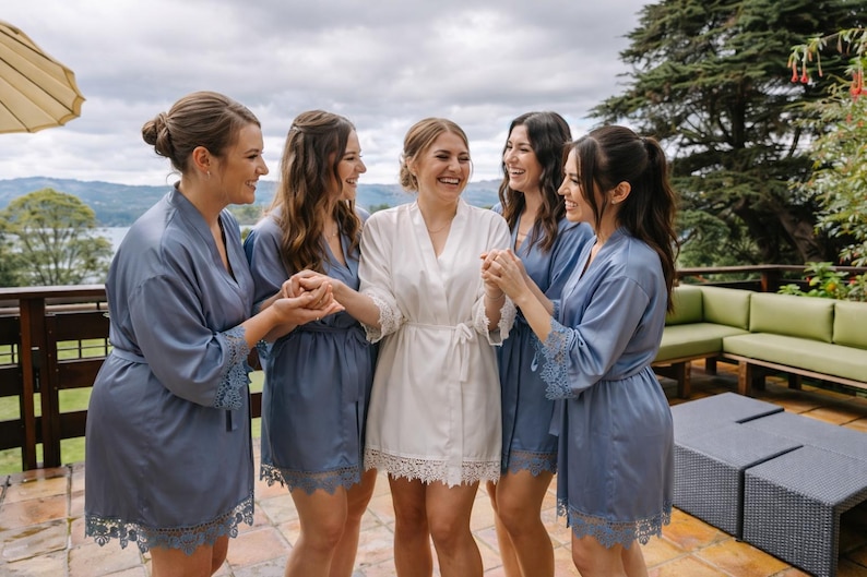 May include: A group of five individuals wearing matching robes. One individual in the center wears a white satin robe with lace trim, while the four surrounding individuals wear dusty blue satin robes with matching lace trim on the sleeves and hem.