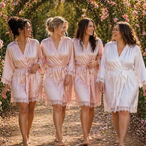 May include: Four robes in shades of light pink and white, each featuring a wrap-style front with a tie waist, long sleeves, and delicate lace trim along the sleeve cuffs and the bottom hem, creating a coordinated set of loungewear garments.