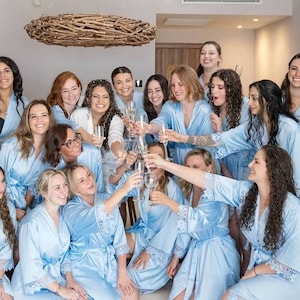 May include: A group of people wearing matching light blue satin robes with white lace trim on the sleeves. The individuals are holding clear glass flutes and raising them in a toast. The robes feature a wrap-style front with a tie waist closure.