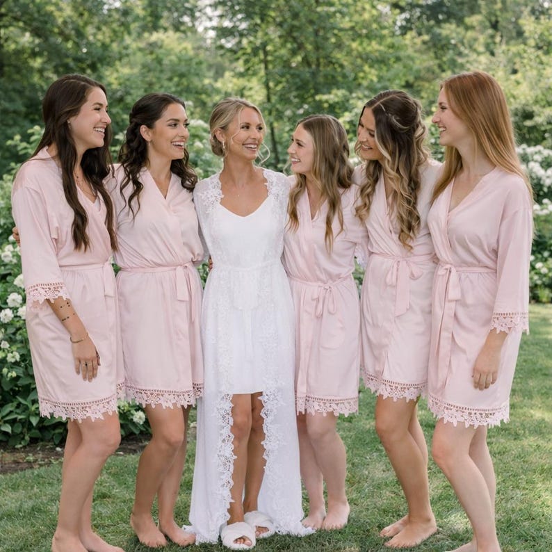 May include: A group of six people, including a bride, are smiling and embracing outdoors. The bride wears a white lace dress, while the others wear matching light pink robes with lace trim. The setting is a garden with green grass and foliage.
