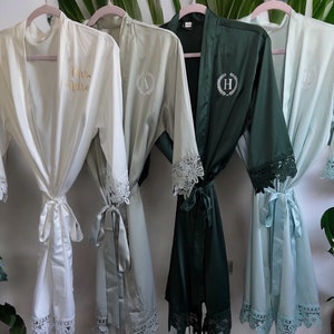 May include: Four satin robes hanging on pink hangers, featuring white, sage green, dark forest green, and light blue colors. Each robe has lace trim on the sleeves and hem, with personalized embroidery on the chest, including letters and script text.