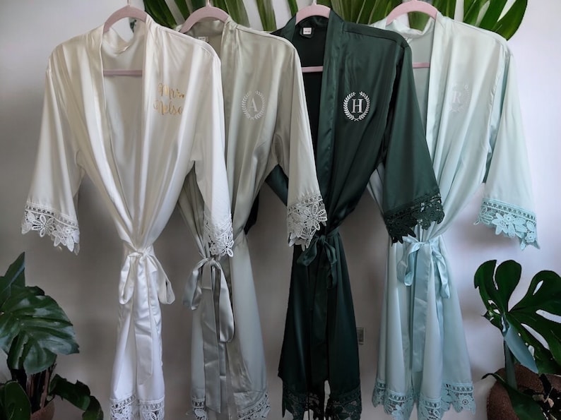 May include: Four satin robes hanging on pink hangers, featuring white, sage green, dark forest green, and light blue colors. Each robe has lace trim on the sleeves and hem, with personalized embroidery on the chest, including letters and script text.