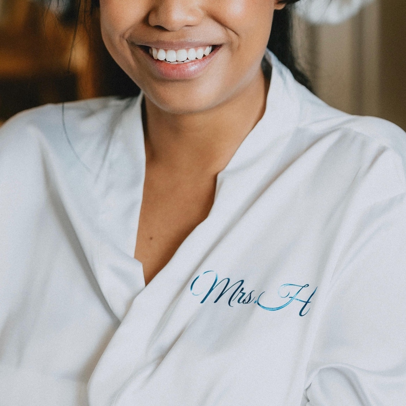 May include: A white satin robe featuring a V-neckline and long sleeves. The left side of the chest is personalized with the text Mrs. H in a metallic blue script font. The fabric has a smooth, slightly reflective finish and a tailored collar.