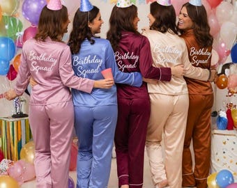 May include: Five individuals wearing long-sleeved satin pajama sets in shades of mauve, blue, burgundy, champagne, and copper. Each shirt features the text Birthday Squad in white script on the back. Each individual wears a colorful party hat.