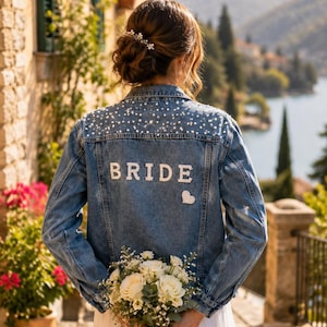 May include: A denim jacket with pearl embellishments and the word "BRIDE" on the back. The jacket is worn over a white dress, and the person is holding a bouquet of white roses. The background features a scenic view.