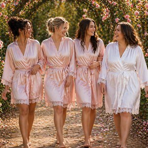 May include: Four matching light pink satin robes featuring long sleeves, a wrap-style front with a tie waist, and delicate white lace trim along the sleeve cuffs and the bottom hemline. The robes have a smooth, shiny finish and a knee-length cut.