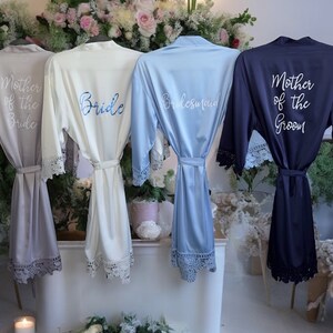 May include: Four satin robes with lace trim on the sleeves and hem. The robes are light gray, white, light blue, and navy blue. They feature white or blue script text reading Mother of the Bride, Bride, Bridesmaid, and Mother of the Groom.