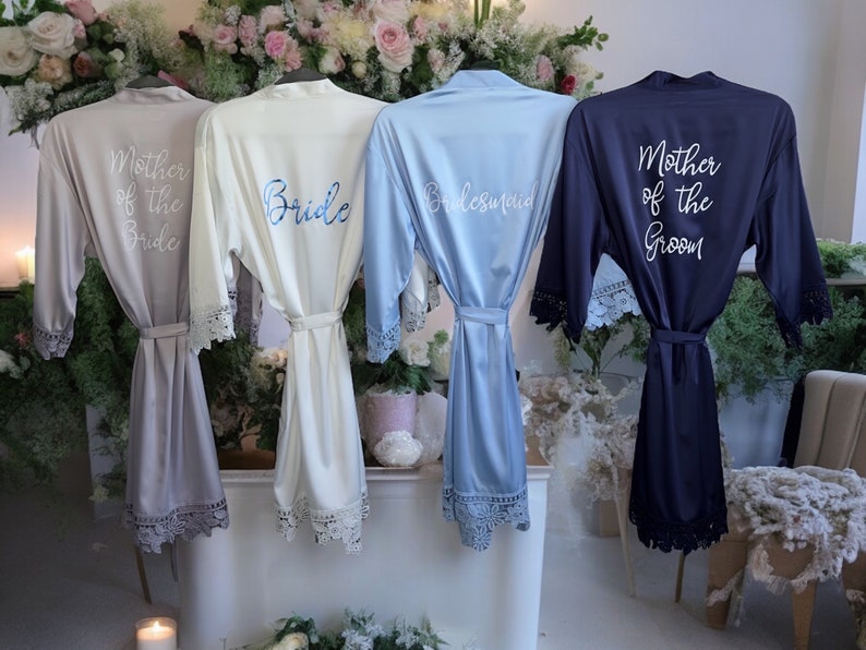 May include: Four satin robes with lace trim on the sleeves and hem. The robes are light gray, white, light blue, and navy blue. They feature white or blue script text reading Mother of the Bride, Bride, Bridesmaid, and Mother of the Groom.