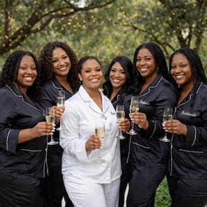 May include: A group of six people, five in black pajamas with white trim and one in white pajamas with gold trim, holding champagne flutes. The pajamas have text on them. The background is a blurred outdoor setting with trees and lights.