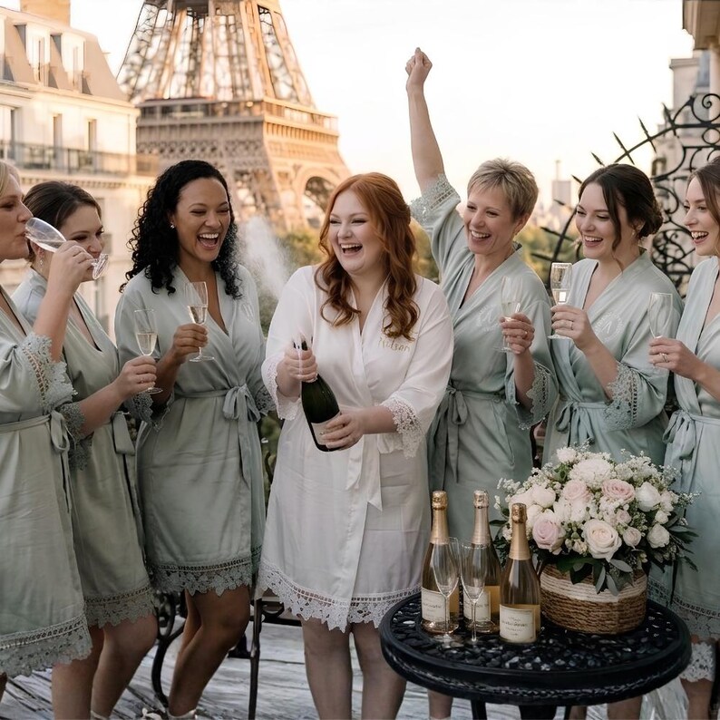 May include: A group of people wearing matching sage green robes and one person in a white robe, all featuring lace trim on the sleeves and hem. The robes have a tie-waist closure and are worn while holding champagne flutes and a bottle of champagne.