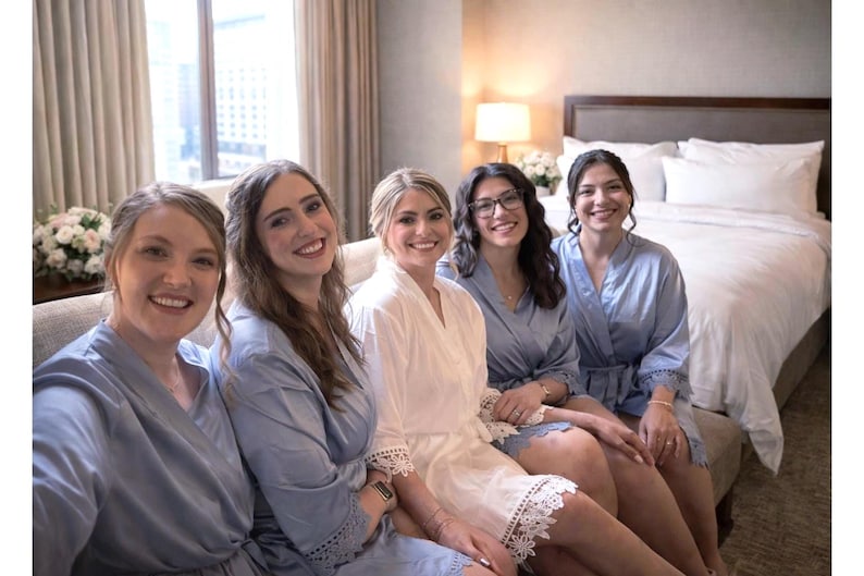 May include: Five women in a well-lit room, smiling at the camera. Four are wearing light blue robes, while one is in a white robe with lace trim. They are seated on a sofa near a window and a bed.