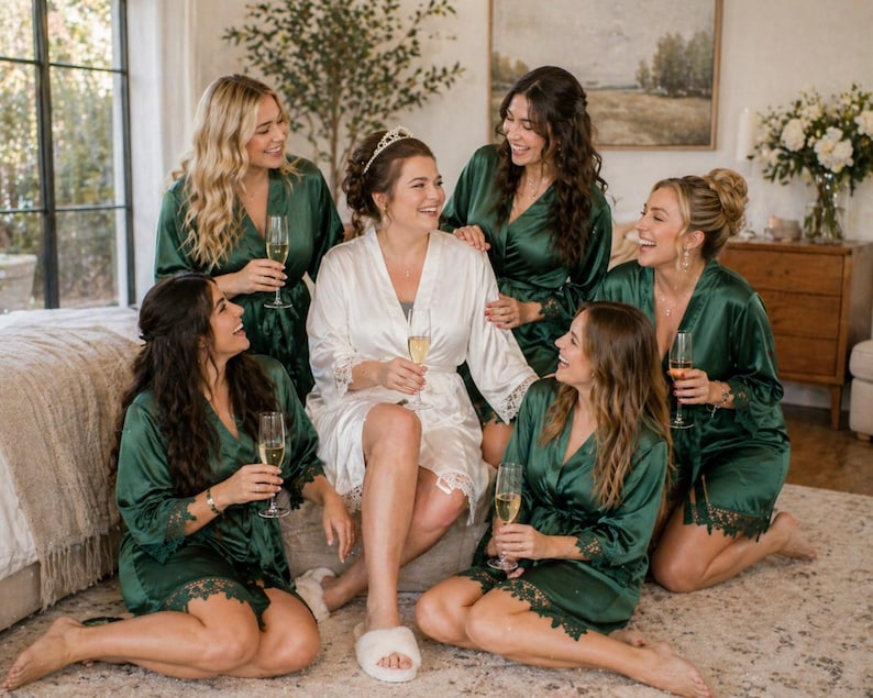 May include: A group of women in emerald green satin robes, some holding champagne flutes, gathered indoors. One woman wears a white robe with lace trim and a tiara. The scene suggests a bridal party or similar celebration.