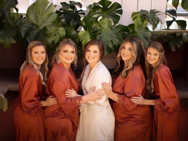 May include: Five women pose for a photo, with a bride in a white robe in the center. The other four women wear rust-colored robes with lace trim. Lush green plants form a backdrop.