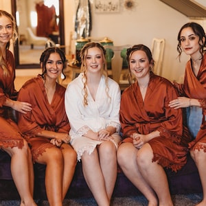 May include: Four individuals wearing knee-length rust-colored satin robes with lace trim on the sleeves and hems, and one individual wearing a knee-length white satin robe with matching lace trim on the sleeves and hems, all seated together.
