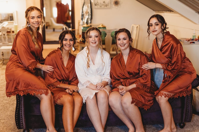 May include: Four individuals wearing knee-length rust-colored satin robes with lace trim on the sleeves and hems, and one individual wearing a knee-length white satin robe with matching lace trim on the sleeves and hems, all seated together.