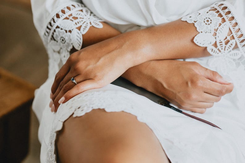 May include: A white robe featuring intricate floral lace trim on the sleeves and hem. A silver ring with a dark blue gemstone is worn on the ring finger. The robe is made of a lightweight fabric and is worn over the lap, with a dark charging cable visible.