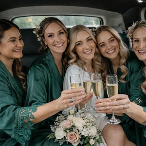 May include: Five smiling women in a car, wearing green robes and holding champagne flutes. One woman wears a white dress and holds a bouquet of flowers. The robes have lace detailing on the sleeves. Floral hair accessories are worn.