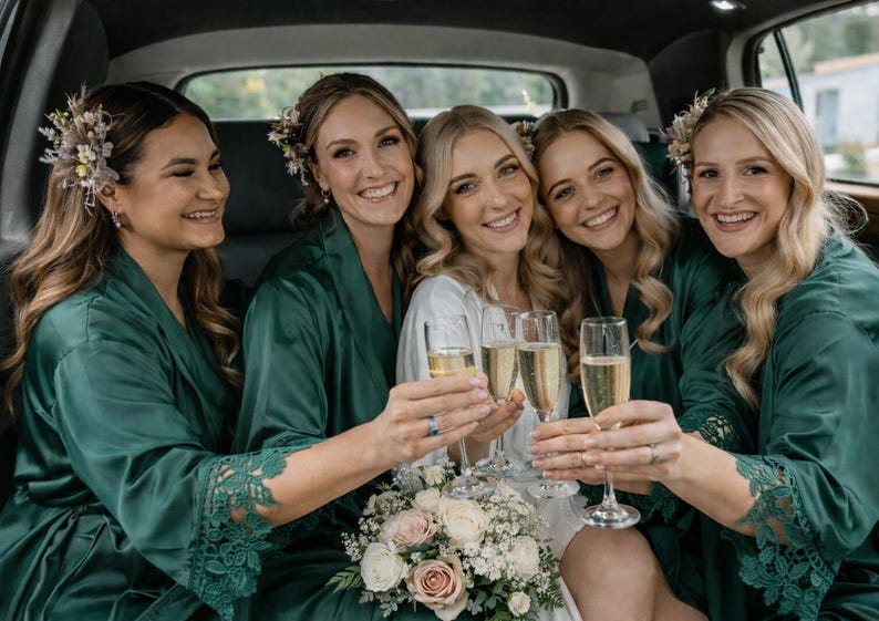 May include: Five smiling women in a car, wearing green robes and holding champagne flutes. One woman wears a white dress and holds a bouquet of flowers. The robes have lace detailing on the sleeves. Floral hair accessories are worn.