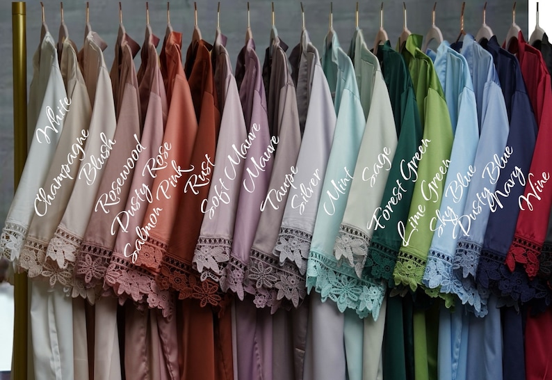 May include: A collection of satin robes hanging on a rack, each featuring intricate lace trim on the sleeves. The robes are labeled with colors including White, Champagne, Blush, Rosewood, Dusty Rose, Salmon Pink, Rust, Soft Mauve, Mauve, Taupe, Silver, Mint, Sage, Forest Green, Lime Green, Sky Blue, Dusty Blue, Navy, and Wine.