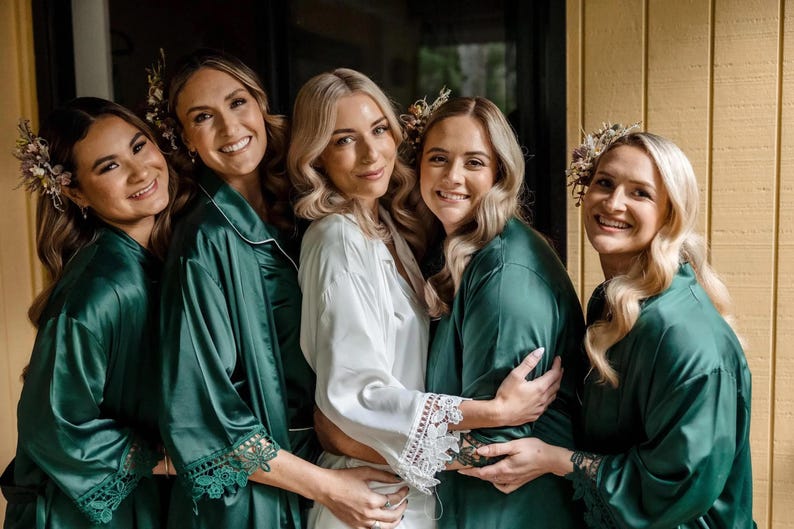 May include: Four individuals wearing emerald green satin robes with lace-trimmed sleeves and one individual wearing a white satin robe with lace-trimmed sleeves. Each robe features a notched collar and a smooth, shiny finish with delicate floral hair accessories.