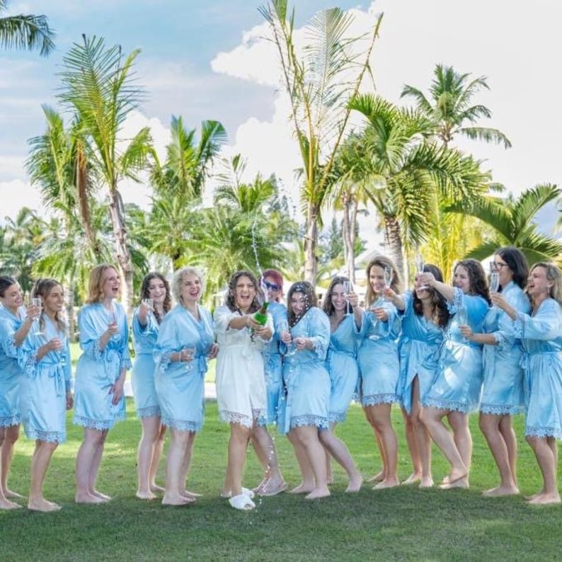 May include: A group of people wearing matching light blue satin robes with lace trim, while one person in the center wears a white satin robe with lace trim. They are standing outdoors in a grassy area with palm trees in the background.