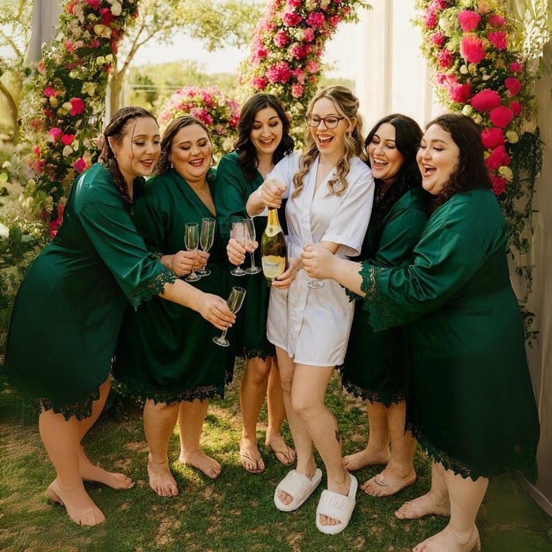 May include: A group of people celebrating, with a champagne bottle being opened. The people are wearing green robes with black lace trim, and one person is wearing a white robe. The background includes a floral arch.