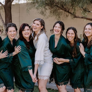 May include: A group of six matching satin robes, five in deep emerald green and one in white, each featuring long sleeves with intricate lace trim at the cuffs and hem. The robes have a wrap-style front with a tie closure and a smooth, shiny finish.