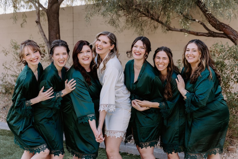May include: A group of six matching satin robes, five in deep emerald green and one in white, each featuring long sleeves with intricate lace trim at the cuffs and hem. The robes have a wrap-style front with a tie closure and a smooth, shiny finish.