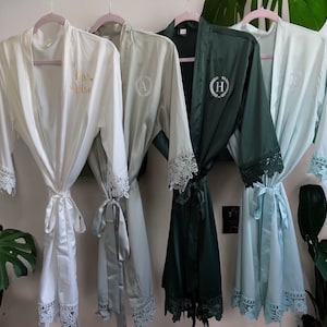 May include: Four satin robes in white, sage green, dark teal, and light blue hang on pink hangers. Each robe has lace trim on the sleeves and a matching tie. The white robe has "Mrs. Nelson" in gold script. Other robes have embroidered initials.