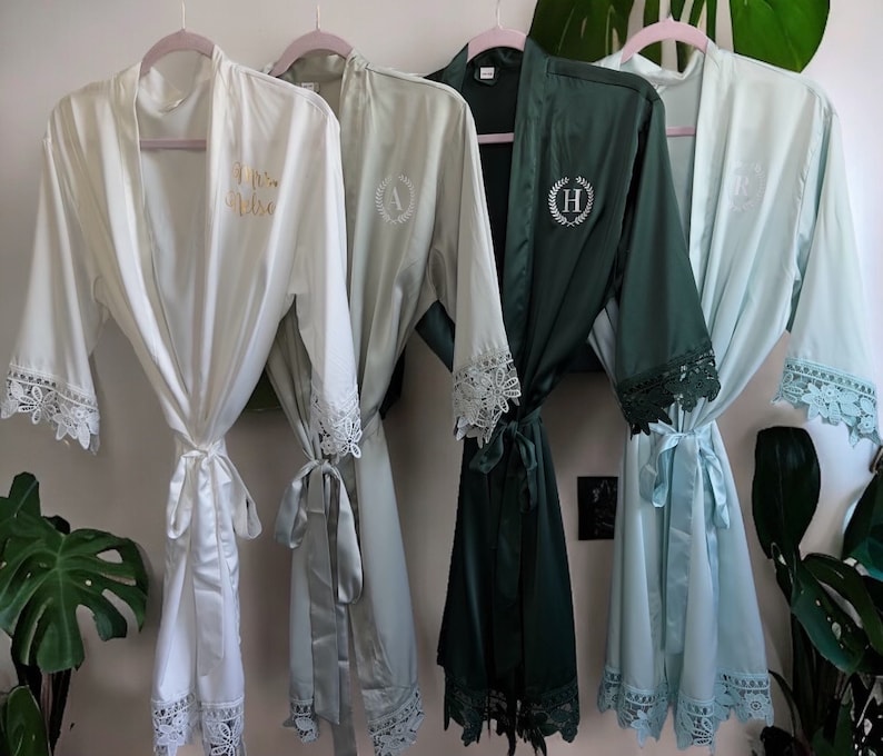 May include: Four satin robes in white, sage green, dark teal, and light blue hang on pink hangers. Each robe has lace trim on the sleeves and a matching tie. The white robe has "Mrs. Nelson" in gold script. Other robes have embroidered initials.