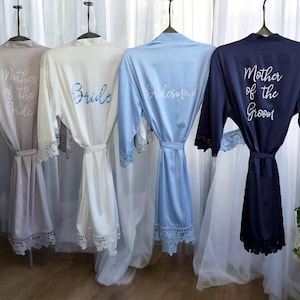 May include: Four satin robes in neutral colors hang on hangers. The robes are embroidered with the words "Mother of the Bride", "Bride", "Bridesmaid", and "Mother of the Groom". Each robe has lace trim on the sleeves and hem.
