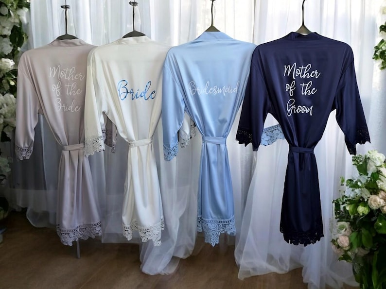 May include: Four satin robes in neutral colors hang on hangers. The robes are embroidered with the words "Mother of the Bride", "Bride", "Bridesmaid", and "Mother of the Groom". Each robe has lace trim on the sleeves and hem.