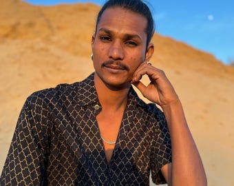 Op de afbeelding: Een persoon met een knot in hun haar draagt een zwart en goud gepatroneerd knoopshirt met een zilveren kettingketting en zilveren armbanden. Ze zitten in een zanderig gebied met hun hand op hun kin.