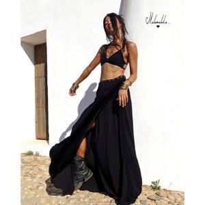 Puede incluir: Una mujer con una falda negra de envoltura con una abertura en la parte delantera y un top de sujetador negro. Lleva pulseras doradas y botas negras. La falda tiene una fila de botones dorados en la parte delantera.