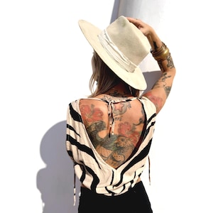 Puede incluir: Un sombrero fedora color crema con ala ancha y cinta crema se levanta. La persona lleva una blusa a rayas negras y crema con la espalda baja. Brazaletes dorados adornan el brazo.