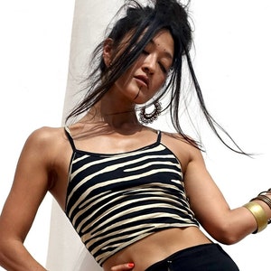 Puede incluir: Un top corto con estampado de cebra en negro y crema con tirantes finos. El top se combina con pantalones negros. La modelo lleva pulseras doradas y un brazalete dorado grande. La modelo también lleva pendientes grandes y ornamentados.