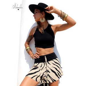 Puede incluir: Una mujer con un top negro, shorts con estampado de cebra y un sombrero negro. Los shorts tienen una cintura negra y un cordón. Pulseras y pendientes dorados completan el look. Fondo blanco.