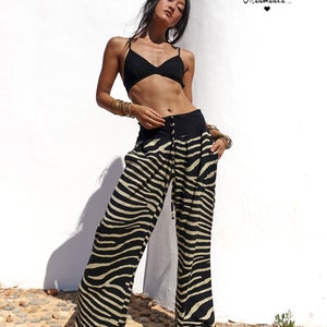 May include: A woman wearing a black bra and zebra print trousers. The trousers have a drawstring waist and wide legs. The woman is standing in front of a white wall with the text "Mahamaka..." written on it.