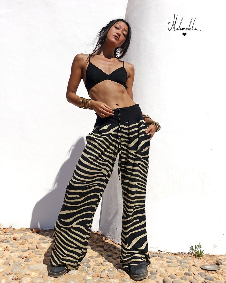 May include: A woman wearing a black bra and zebra print trousers. The trousers have a drawstring waist and wide legs. The woman is standing in front of a white wall with the text "Mahamaka..." written on it.