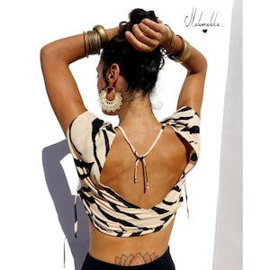 Puede incluir: Una blusa crema con la espalda abierta y un estampado de rayas de cebra negras. La blusa tiene mangas cortas y cierres de lazo. La modelo lleva pendientes de aro y pulseras doradas. La imagen incluye el texto "Mahamalaka..."