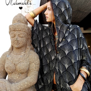 May include: Black hooded cape with a fan-like pattern in white. The cape has a button-down front and pockets. The image also shows a stone statue and the text "Malamalaka..." in cursive.
