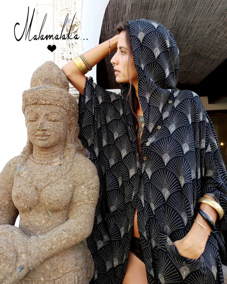 May include: Black hooded cape with a fan-like pattern in white. The cape has a button-down front and pockets. The image also shows a stone statue and the text "Malamalaka..." in cursive.