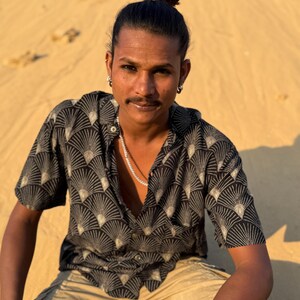 Op de afbeelding: Een zwart overhemd met een waaierachtig patroon in beige en wit. Het shirt met korte mouwen wordt gedragen met een beige broek. De persoon draagt een zilveren ketting en oorbellen.