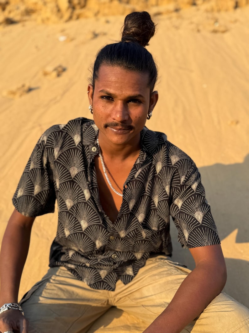 Op de afbeelding: Een zwart overhemd met een waaierachtig patroon in beige en wit. Het shirt met korte mouwen wordt gedragen met een beige broek. De persoon draagt een zilveren ketting en oorbellen.